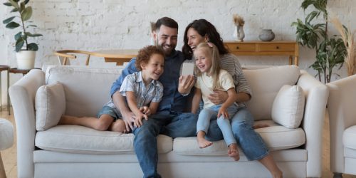 Happy excited couple of parents, little son and daughter kids talking on video call, taking selfie on cellphone, chatting, smiling, laughing, having fun, hugging on comfortable sofa at home